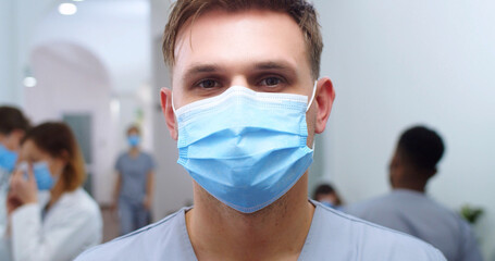 Close up portrait of handsome young Caucasian male doctor in medical mask standing in hospital and looking at camera. Man surgeon at work in medical center in coronavirus. Epidemic concept