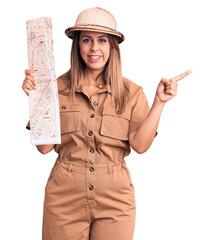 Young beautiful woman wearing explorer hat holding map smiling happy pointing with hand and finger...