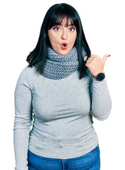 Young hispanic plus size woman wearing winter scarf surprised pointing with hand finger to the side, open mouth amazed expression.