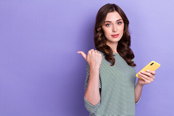 Photo of young serious lady wear striped shirt nice wavy hair finger directing empty space channel hold phone isolated on purple color background