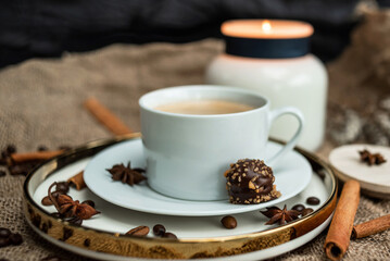 An aesthetic composition of a cup of black coffee with foam and a burning candle, decorated with chocolates, anise stars, coffee beans and cinnamon sticks