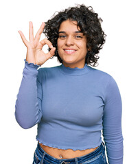 Young hispanic woman with curly hair wearing casual clothes smiling positive doing ok sign with hand and fingers. successful expression.