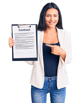 Young beautiful latin girl holding clipboard with contract document smiling happy pointing with hand and finger