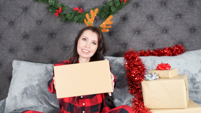Happy young woman wearing red flannel pajama with holiday deer antlers and holding gift box with space for text sitting on bed with Christmas decorations, presents, copy space. Selective focus.