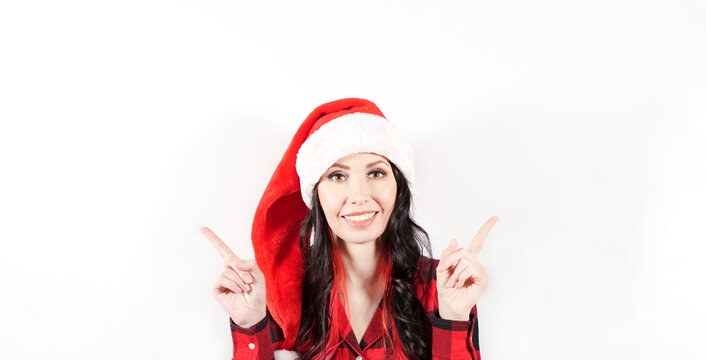 Smiling woman in red Christmas hat holding finger at empty Space for text for promo discount. Beautiful lady, Long curly hair, red santa hat, red pajama. Woman portrait isolated on white background.
