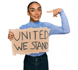 Hispanic transgender man wearing make up and long hair holding united we stand banner pointing finger to one self smiling happy and proud