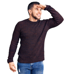 Young handsome man wearing casual sweater very happy and smiling looking far away with hand over head. searching concept.