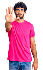 Handsome young man with curly hair and bear wearing casual pink tshirt doing stop sing with palm of the hand. warning expression with negative and serious gesture on the face.