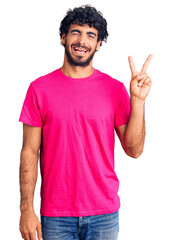 Naklejka premium Handsome young man with curly hair and bear wearing casual pink tshirt smiling with happy face winking at the camera doing victory sign. number two.