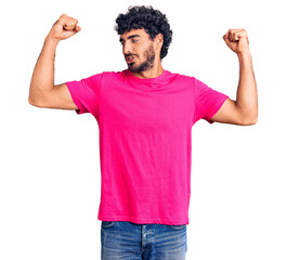 Handsome young man with curly hair and bear wearing casual pink tshirt showing arms muscles smiling proud. fitness concept.
