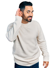 Young hispanic man with beard wearing casual white sweater smiling with hand over ear listening an hearing to rumor or gossip. deafness concept.