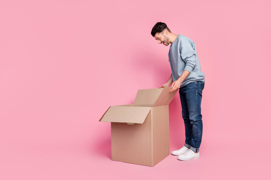 Full length photo of attractive young man bending over peek inside carton box wear stylish blue garment isolated on pink color background