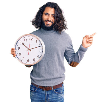 Young arab man holding big clock smiling happy pointing with hand and finger to the side