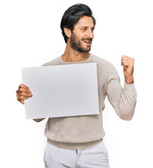 Young hispanic man holding blank empty banner pointing thumb up to the side smiling happy with open mouth