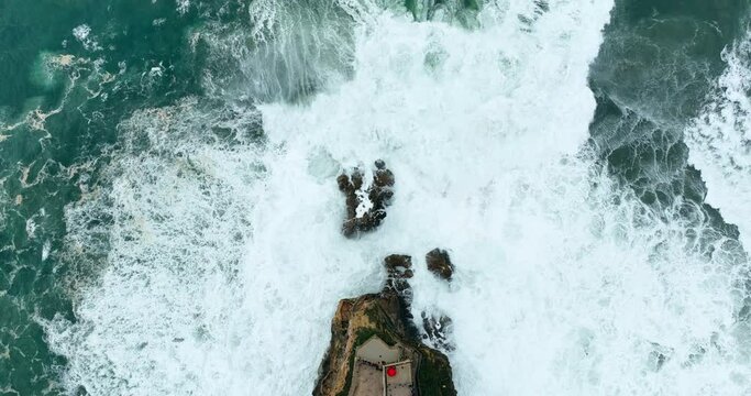 Aerial view of a cliff with a fortress on the coast of the Atlantic ocean. A big rocky cliff with a lighthouse on it. Huge waves at storm in Nazare - famous surfing spot in Portugal.