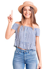 Young beautiful girl wearing hat and t shirt showing and pointing up with finger number one while smiling confident and happy.