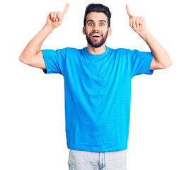 Young handsome man with beard wearing casual t-shirt smiling amazed and surprised and pointing up with fingers and raised arms.