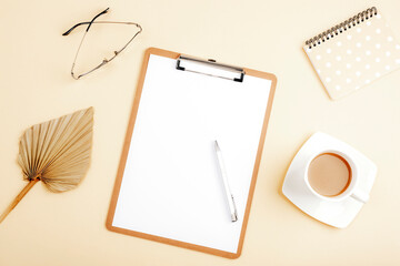 Desktop with blank clipboard, notepad, fan leaf, eyeglasses and accessories on light beige background. Top view, flat lay, mockup