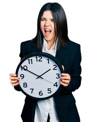 Young brunette woman with blue eyes holding big clock angry and mad screaming frustrated and furious, shouting with anger. rage and aggressive concept.