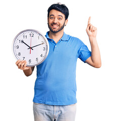 Young hispanic man holding big clock surprised with an idea or question pointing finger with happy...