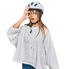 Teenager caucasian girl wearing bike helmet doing ok gesture shocked with surprised face, eye looking through fingers. unbelieving expression.