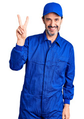 Middle age handsome man wearing mechanic uniform smiling looking to the camera showing fingers doing victory sign. number two.