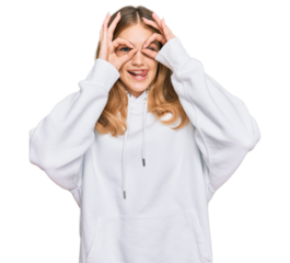 Beautiful young caucasian girl wearing casual sweatshirt doing ok gesture like binoculars sticking tongue out, eyes looking through fingers. crazy expression.
