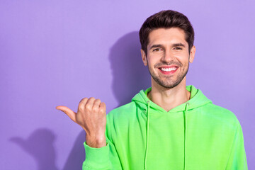 Photo of positive handsome nice guy with brunet hairdo dressed green hoodie indicating empty space isolated on purple color background