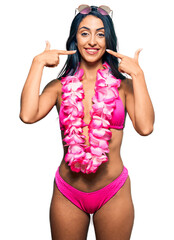Beautiful hispanic woman wearing bikini and hawaiian lei smiling cheerful showing and pointing with...