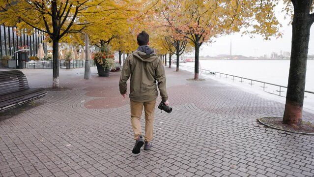 Young male photographer in outdoor clothing walking through an urban city park searching for photo opportunities with camera in hand