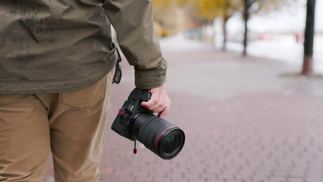 Close up of photographer walking outdoors during the Fall season holding a professional camera in hand. Walks away from camera searching for photo opportunities
