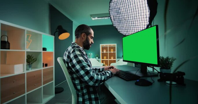 Young freelancer, copywriter, designer, editor or content creator working on desktop computer with chroma key green screen mock up display. Modern office studio with teal and orange lights. Wide shot.