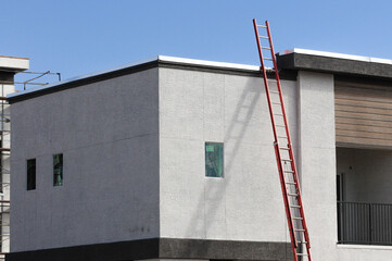 Corner of a modern residential building under construction with a red extension ladder against the stucco wall, illustrating exterior finishing and roof access in housing development. © Eduardo Barraza