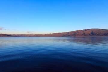 青空の下で遠くの山が朝陽にオレンジ色に染まる湖の風景。