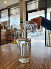 A woman pours mineral water from a plastic bottle into a glass almost full. Glass of water on wooden table on blurred background. Drink about 2 liters of water per day to compensate for the water lost