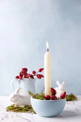 Foto auf Leinwand White burning candle, moss, briar berries and white bunnies on white marble background against blue  textured wall. Rabbit is symbool of 2023.. Selective focus. Place for text. © daffodilred