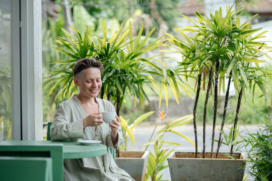 Beautiful woman on the terrace of the cafeteria enjoys a delicious coffee drink among the green foliage.