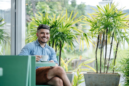 Positive confident man sits on a terrace in tropical greenery and drinks a coffee drink.