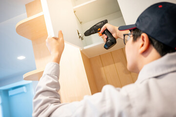 Asian male furniture assembler using electric drill screwdriver on stainless steel cabinet hinge....