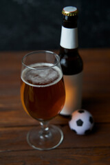 Two glasses with beer, bottles, snacks and soccer ball on wooden table.
