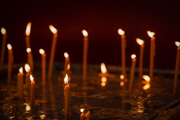 Unfocused melting wax thin candles with small flames stand on church candle table in dark, only the...