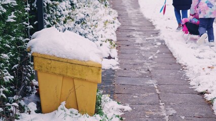 Yellow Plastic Winter Grit Sand Box Covered in Snow Standing by Icy Pavement