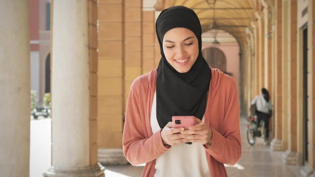 happy pleased muslim girl using smart phone mobile walking along city street,arab young woman typing on smartphone outdoor