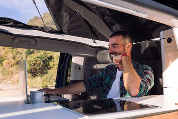 smiling young man working with his laptop sitting in his camper van, concept of freedom and digital nomad lifestyle