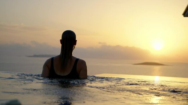 slender unrecognizable young woman emerges from infinity pool with beautiful view of sea and sunset. Silhouette of girl get out of water in pool with the ocean and sunrise in background.