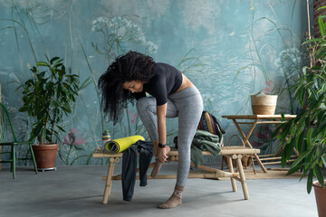 Curly African-American woman putting on clothes after fitness training over green domestic house plant loft blue home background. Lifestyle concept. Copy space, mock-up. Eco natural, body conscious - obrazy, fototapety, plakaty