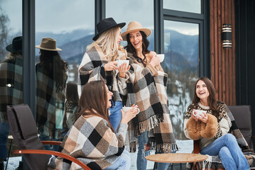 Young women enjoying winter weekends on terrace of contemporary barn house. Four girls having fun, sitting on chairs and drinking hot tea.