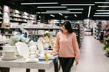 A young brunette woman in a pink sweater buys tableware in a store.