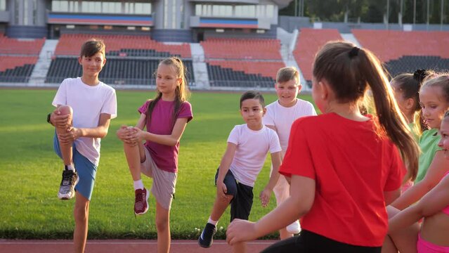 A group of children, on the treadmill at the stadium on a bright sunny day, warm up with the coach before the competition. Boys and a girl in colorful uniforms at the stadium. A sporty lifestyle. 