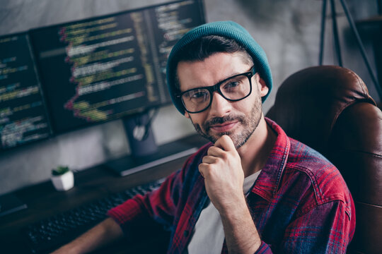 Photo of positive thoughtful coder dressed hat glasses smiling arm chin game developing indoors workplace workstation loft